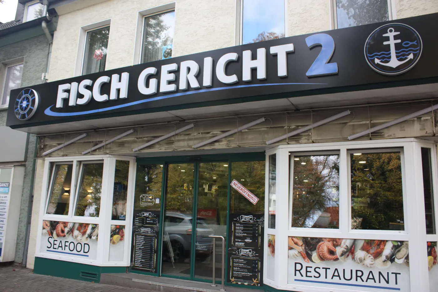 Der Laden von außen: schwarzes Schild Fisch Gericht 2 über dem Eingang, Schaufenster mit Seafood und Restaurant